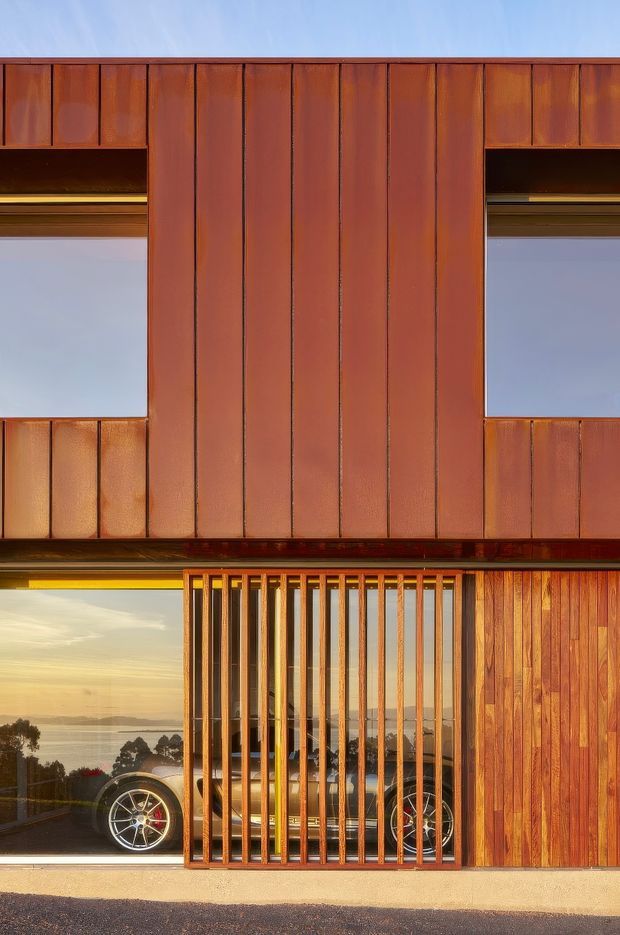 Exterior architectural view of a contemporary multi-level building clad in richly weathered reddish-brown vertical metal panels and timber. Large glazed openings reveal a garage with a sports car, while a prominent vertical timber screen forms a gated frontage. A calm coastal or waterfront landscape is visible through the glazing, with strong late-afternoon light.