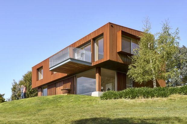 Exterior view of a modern two-storey home set into a landscaped grassy site. The building has a strong rectilinear form, extensive glazing, warm vertical timber cladding, a projecting upper-level balcony, and a partially cantilevered upper volume. Mature planting screens part of the facade.