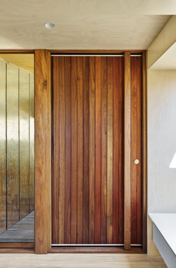 Interior architectural detail focused on a tall vertical timber sliding or pivot door with rich dark wood battens and matching timber framing. A textured glass partition and reflected timber flooring are visible to the left, while a pale built-in surface and light wood ceiling and wall finishes frame the composition. The image has a warm, refined mid-century-inspired material palette and strong emphasis on joinery and natural wood grain.