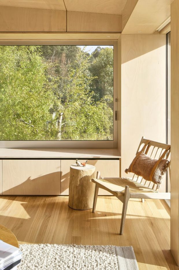 A warm, sunlit minimalist interior with pale timber wall panelling, built-in cabinetry and a large picture window overlooking dense greenery. A timber lounge chair with a woven seat, a simple tree-trunk side table, and a textured light rug create a calm reading corner. The room features a light timber floor, strong natural light and restrained natural materials.