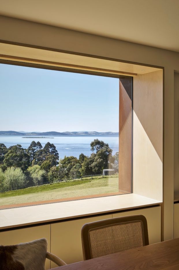 Interior view of a warm, wood-lined dining or workspace nook positioned beside a large picture window. The deep timber window reveal includes a built-in sill or desk surface, with a wooden table and woven-back chair in the foreground. The window frames a bright waterfront landscape with trees, lawn, calm water and distant hills.