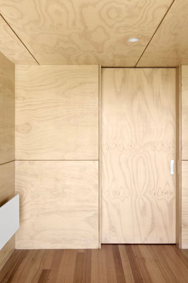 A minimalist interior hallway or entry area lined with pale plywood wall and ceiling panels. A flush plywood door with a slim white handle is centered in the frame, with matching built-in panel detailing and warm timber plank flooring. A recessed ceiling light provides subtle illumination. The design is simple, warm, and highly cohesive through the extensive use of natural timber.