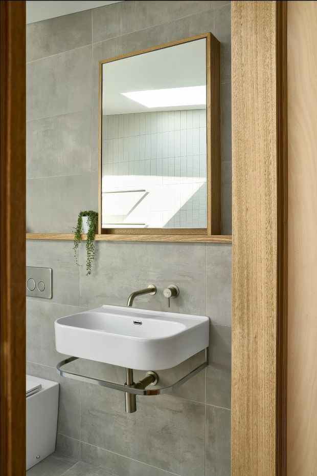 A compact contemporary bathroom viewed through a timber-framed doorway. The room features large grey concrete-look wall and floor tiles, a wall-mounted white ceramic basin with exposed chrome/silver support frame and plumbing, brushed brass-toned wall-mounted tapware, a rectangular timber-framed mirror, a dual-flush plate, and a narrow timber ledge with a small trailing plant. Bright natural light is reflected in the mirror from a skylight or roof opening.
