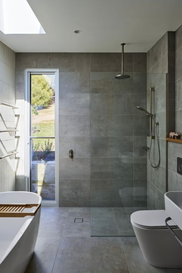 Contemporary bathroom with a walk-in glass shower, large grey stone-look wall and floor tiles, a freestanding white bathtub, wall-hung toilet, floating vanity, and dark metal shower fittings. A skylight and tall window provide abundant natural light, while a narrow timber shelf adds warmth.