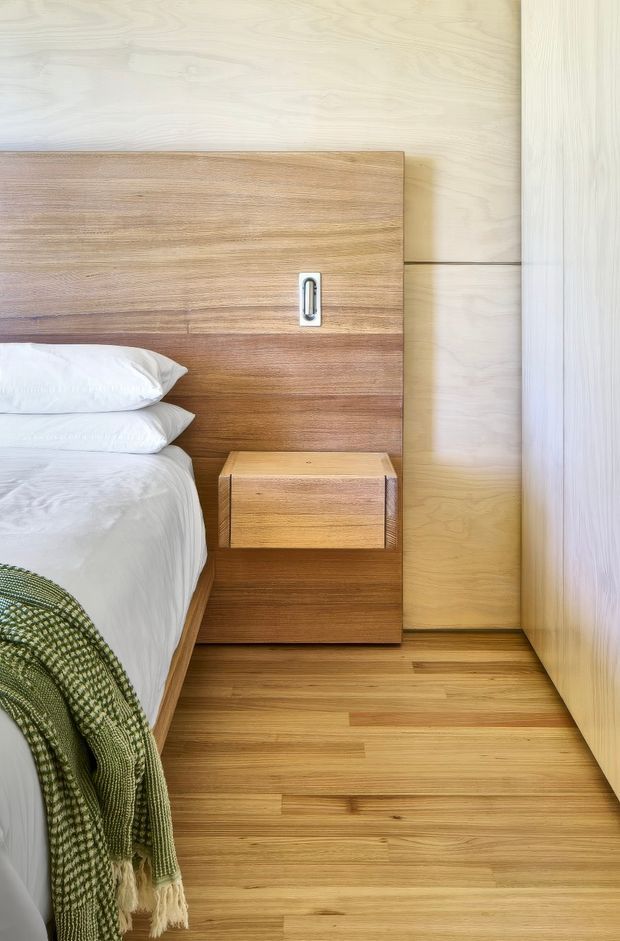 Close-up view of a compact, warm-toned bedroom featuring a bed with white linens and a textured green throw, a built-in timber headboard with integrated bedside storage, pale timber wall panelling, a timber floor, and a tall pale timber wardrobe or cabinet. The design is simple, functional, and strongly coordinated through natural wood finishes.