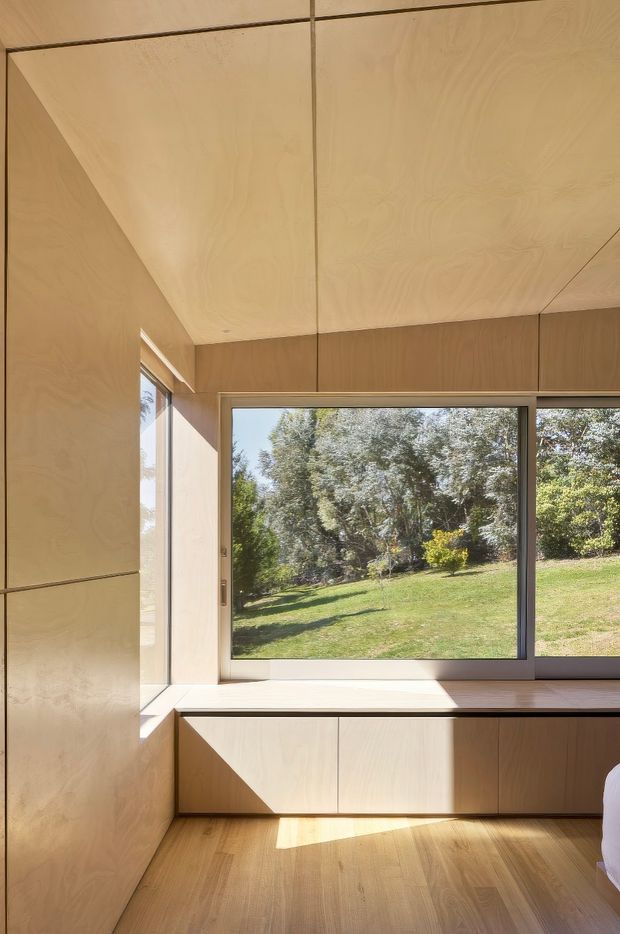 A compact, light-filled room lined extensively with pale plywood panels on the walls, ceiling, built-in window seat, and cabinetry. A large horizontal window overlooks a leafy lawn and trees, while warm timber flooring and strong sunlight create a calm, minimal interior. The edge of a bed is visible at the lower right, indicating a bedroom.