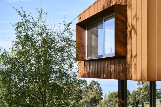Close-up exterior view of a contemporary elevated building clad in warm brown vertical timber boards. A projecting box window with dark framing and reflective glazing interrupts the facade, while slender dark supports lift the upper volume above a leafy landscape. The image emphasizes the building envelope, window projection, and integration with trees and open sky.