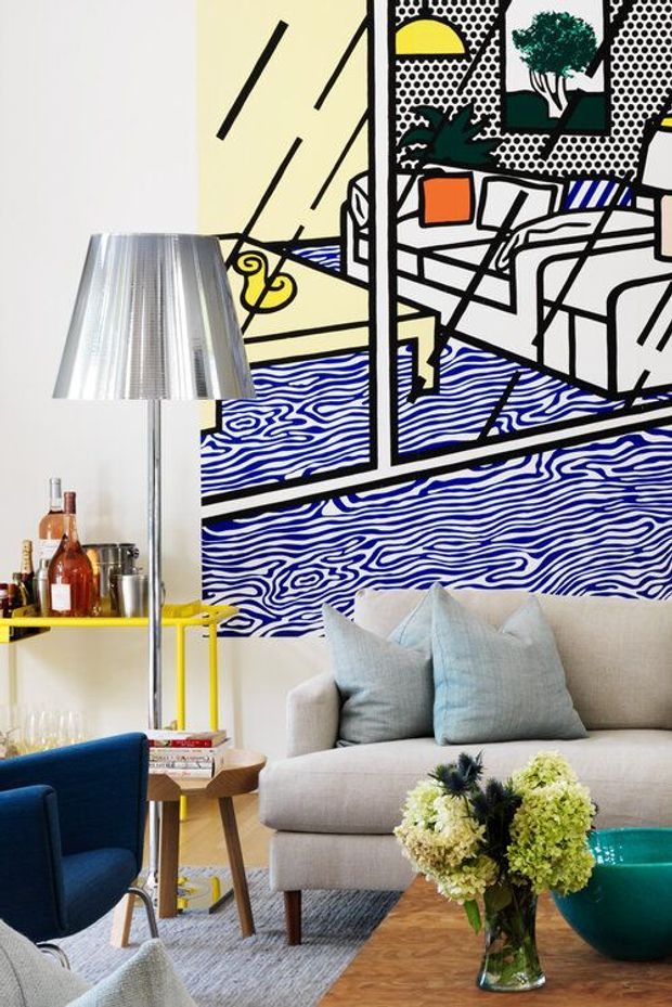 A colourful contemporary living room featuring a large graphic black, white, yellow and blue wall artwork, a light beige sofa with pale blue cushions, a deep blue armchair, a yellow round drinks trolley, and a teal side table. A metallic floor lamp, timber furniture, patterned rug and floral arrangement complete the eclectic interior.