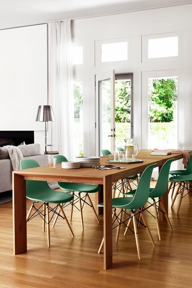 Bright open-plan dining and living area with a large solid timber dining table surrounded by green molded chairs, set on warm timber flooring. White walls, full-height curtains, extensive white-framed glazing and French doors bring in abundant natural light. A partially visible fireplace and sofa sit to the left, with tableware and a pendant lamp completing the dining setting.