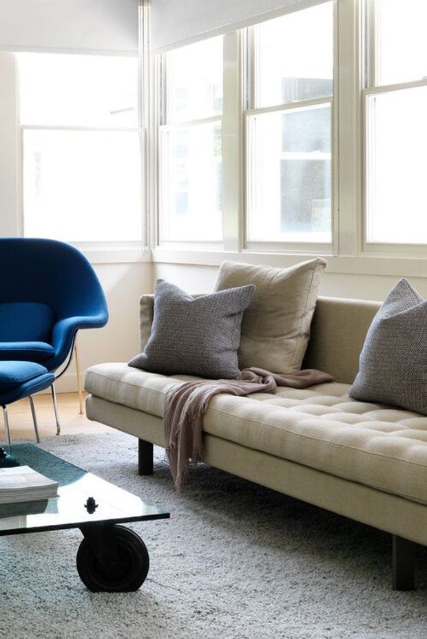 Bright living room with large white-framed windows, a cream tufted sofa, a vivid blue sculptural armchair, neutral cushions, a soft grey rug, and a low glass coffee table with a dark sculptural base. The room has a clean, refined mid-century-inspired aesthetic with abundant natural light.
