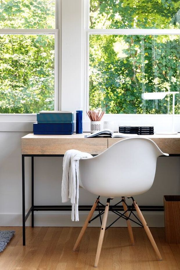 Bright, compact home office with a simple timber-topped desk, white molded chair with wooden legs, desk lamp, books, stationery, and storage boxes. Large white-framed windows overlook leafy greenery, while timber flooring and minimal furnishings create a clean, airy workspace.