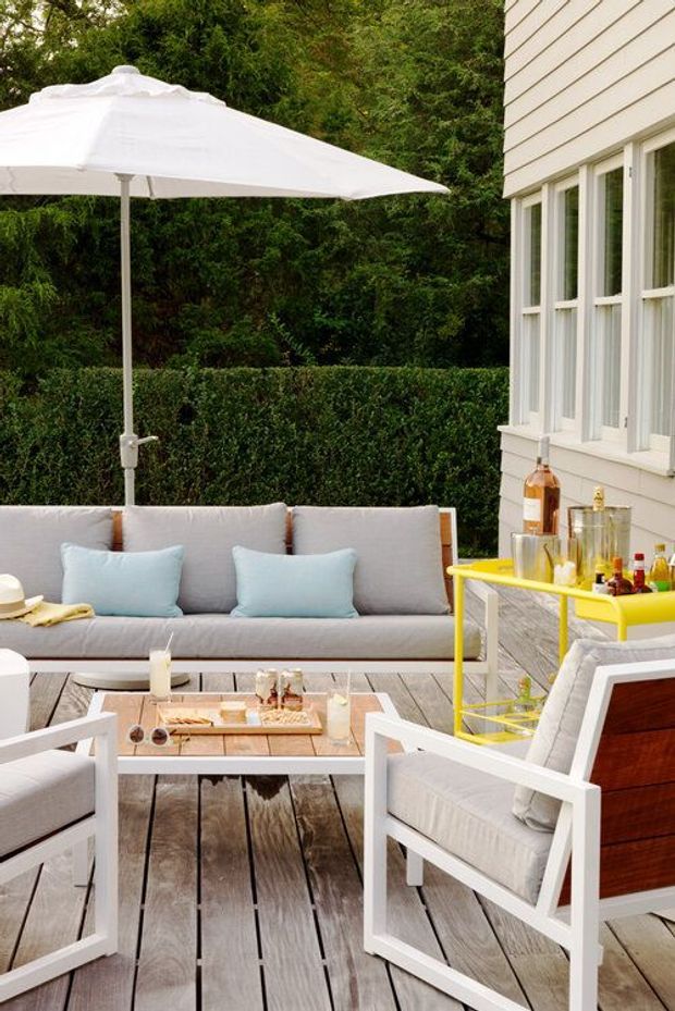 A stylish outdoor lounge on a timber deck beside a white weatherboard-clad house. The space features a large white umbrella, light grey cushioned seating with pale blue pillows, a low coffee table, and a bright yellow drinks trolley stocked for entertaining. Dense greenery and clipped hedges provide a private garden backdrop.