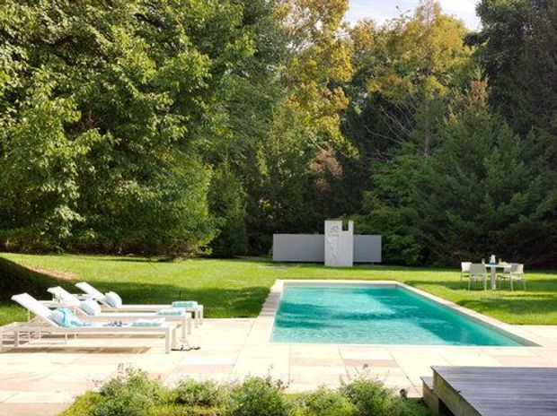 A landscaped outdoor swimming pool area with a rectangular turquoise pool, pale paved surround, white sun loungers, and a built-in outdoor bar or counter at the far end. The pool is set within a large manicured lawn surrounded by mature trees and dense greenery, with a timber deck visible in the foreground.