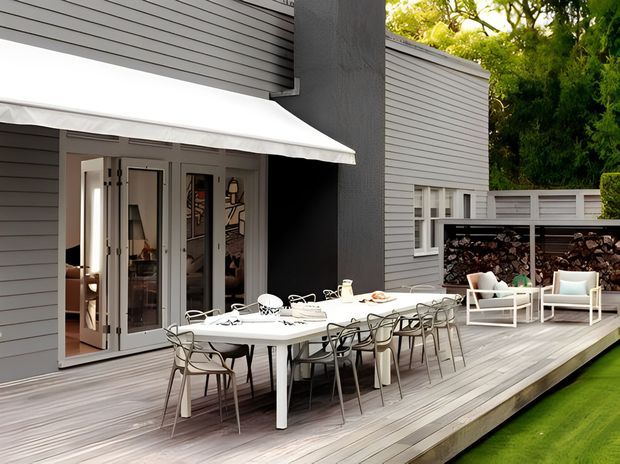 A contemporary outdoor entertaining deck beside a grey horizontal-clad house. The spacious timber deck features a long outdoor dining table with numerous chairs, a separate lounge seating area, built-in firewood storage, a white awning, and folding glazed doors opening into the interior. Lush greenery and lawn frame the scene.