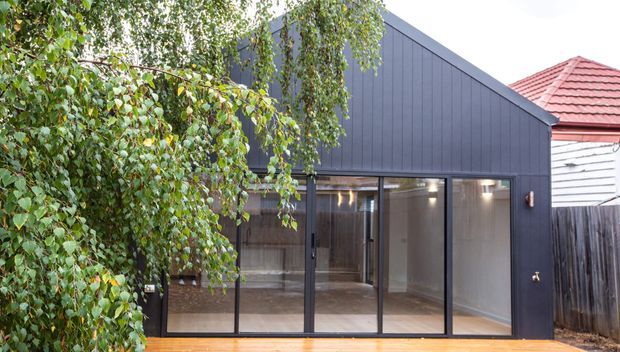 Exterior view of a compact modern addition with dark vertical cladding, a steep mono-pitched roof, and a full-width glazed wall with sliding or hinged glass doors. A timber deck runs along the front, with mature planting screening the left side and a neighbouring traditional red-tiled roof visible behind.