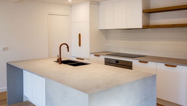 A bright contemporary kitchen featuring a large pale stone island with an integrated black sink and bronze-toned gooseneck tap. White flat-panel cabinetry with warm wood handles surrounds a built-in cooktop and oven, while a fine vertical tile backsplash and open timber shelves add texture and warmth.