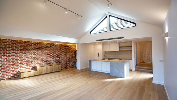 Bright open-plan living and kitchen area with a high vaulted ceiling, exposed red brick feature wall, light timber flooring, white cabinetry, and a central island with a pale stone benchtop. Clerestory-style triangular glazing brings in natural light, while a timber door and short stairway are visible at the rear.