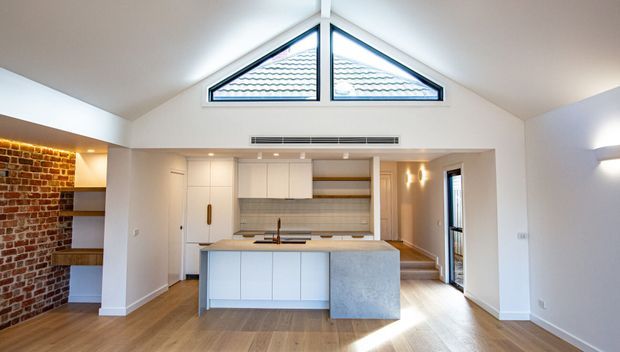 Bright open-plan kitchen and living area with a vaulted ceiling and prominent triangular clerestory windows. The kitchen features a large white island with a grey stone waterfall end, white cabinetry, timber flooring, warm under-cabinet lighting, and an exposed red-brick feature wall with open shelving.