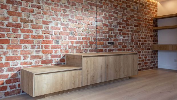 A warm contemporary interior featuring an exposed red brick feature wall, light timber flooring, and minimalist built-in timber cabinetry. The cabinetry includes a low floating unit and a taller storage unit, with open timber shelves recessed into the wall at right. The space appears to be a living or media area, though no seating or screen is visible.