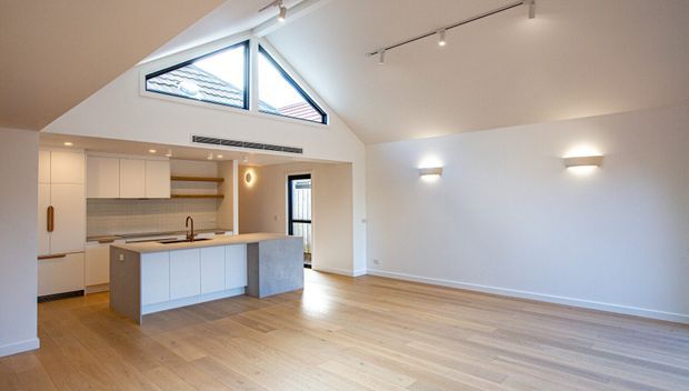 Bright open-plan interior combining a modern white kitchen with island and an adjacent spacious living area. The room has light timber flooring, white walls and cabinetry, a high sloping ceiling, triangular clerestory windows, and contemporary track and wall lighting.