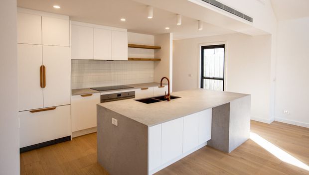 Bright contemporary kitchen with a large concrete-look island, white integrated cabinetry, open timber shelves, fine grid-tile splashback, timber flooring, and black-framed glazing. Recessed ceiling lighting and a linear air-conditioning vent contribute to the clean, minimalist fit-out.