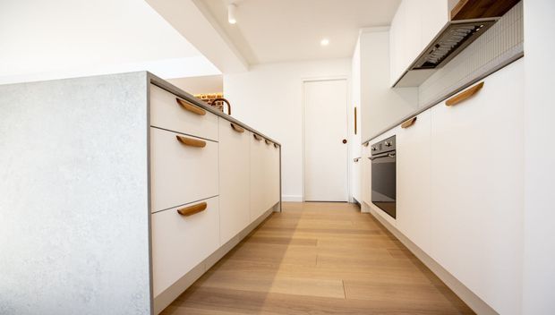 Bright, compact contemporary galley kitchen with white flat-panel cabinetry, warm timber pulls, integrated oven and cooktop, a pale stone island with drawers, and light timber flooring. Recessed ceiling lights and a white door at the rear create a clean, efficient layout.