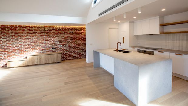 Bright open-plan kitchen and living area with a large pale stone island, white flat-panel cabinetry, integrated appliances, open timber shelving, and a textured white tiled splashback. The adjacent living zone features a striking exposed red-brick feature wall, low timber joinery, and light timber flooring. High-level glazing and recessed lighting enhance the airy contemporary interior.