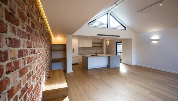 Open-plan contemporary living area with a compact white kitchen and island, light timber flooring, exposed red brick feature wall, and a pitched ceiling with triangular clerestory glazing. Built-in timber shelving and stepped seating are integrated along the brick wall, while track lighting and concealed linear lighting provide illumination.