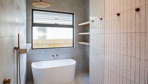 A compact contemporary bathroom featuring a freestanding white bathtub, grey tiled walls and floor, a black-framed frosted window, open wall shelving, and a vertically ribbed pale tile feature wall with dark wall-mounted hooks. A round ceiling shower outlet is visible above.