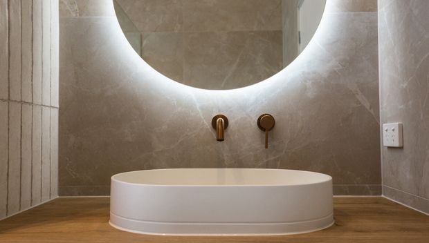 A refined bathroom featuring a freestanding oval white bathtub set against full-height warm beige stone-look wall tiles. A large round backlit mirror is mounted above the bath, with minimalist brushed brass wall-mounted tapware and a timber-look floor. Vertical fluted wall detailing appears at the left edge, creating a calm, softly illuminated atmosphere.