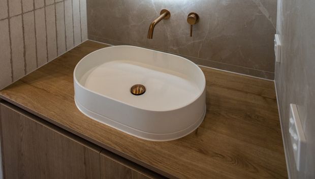 Close-up of a contemporary bathroom vanity featuring a white oval vessel basin on a warm wood countertop, with a wall-mounted brass/gold faucet and controls. The surrounding walls use large neutral stone-look tiles, while the vanity cabinetry below has a wood-grain finish.