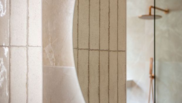 Close-up view of a bathroom shower area featuring pale textured and vertically grooved ceramic tile surfaces, a curved tiled partition or wall edge, and brushed copper or bronze shower fittings visible through a glass screen. The palette is warm, soft, and neutral with subtle material variation.