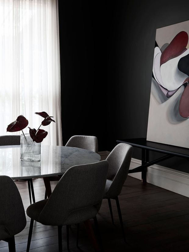 A sophisticated dining area with a round pale marble dining table surrounded by dark upholstered dining chairs. The room features dark charcoal walls, dark timber flooring, sheer floor-to-ceiling curtains, a slim black console table, and a large abstract artwork with burgundy, black, white, and grey forms. The space has a moody, refined contemporary feel with strong natural light filtering through the curtains.