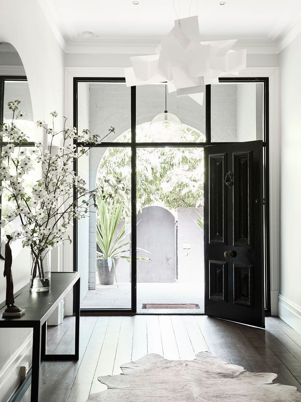 Bright, refined entryway with white walls, ornate ceiling cornices, pale timber floorboards and a dramatic black framed glazed front door. A black console table with botanical branches sits to the left, while a sculptural white pendant hangs centrally. The entry opens onto a small outdoor courtyard visible through the glazing.