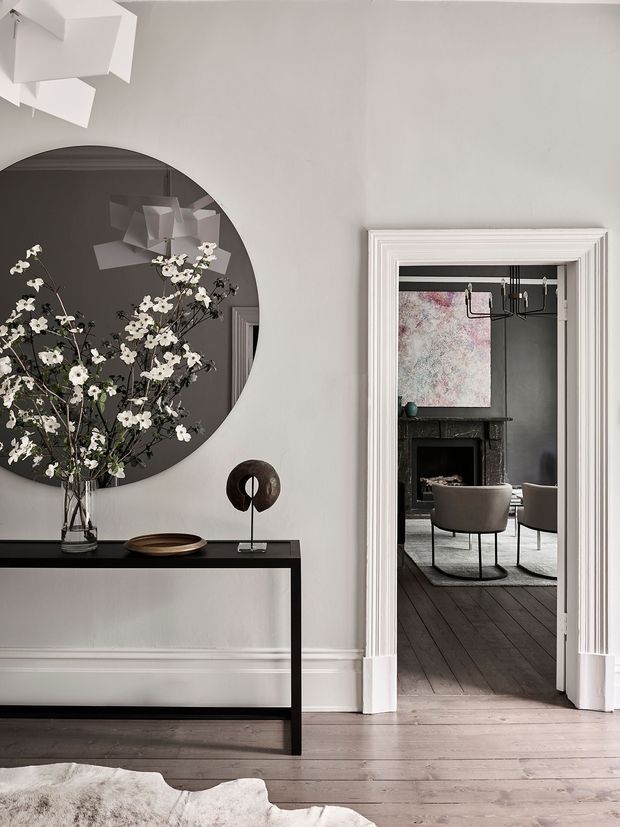 Elegant residential interior showing a refined hallway or entry space opening into a living room. The foreground features a slim black console table, sculptural decor, a large round dark mirror, and a tall floral arrangement. White walls, ornate layered architraves, decorative skirting, and pale timber floorboards create a classic architectural shell, while the adjoining room has dark timber flooring, a fireplace, upholstered lounge chairs, a chandelier, and abstract artwork. The palette is restrained in white, grey, black, and muted timber tones with a sophisticated, gallery-like atmosphere.