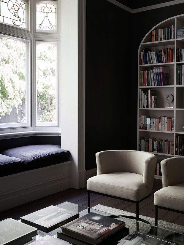 A refined living room with a dark painted wall, built-in arched bookshelves filled with books, a bay-window reading bench with a dark cushion, and two light upholstered lounge chairs around a glass coffee table. Decorative stained-glass upper window panels, detailed white trim, and dark timber flooring create a classic, sophisticated atmosphere.