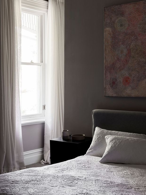 A moody, refined bedroom interior with a dark charcoal wall, upholstered bedhead, black bedside table, and layered white textured bedding. Full-height sheer curtains frame a tall white-trimmed window, while a muted abstract artwork hangs above the bed. Soft daylight creates strong contrast between the bright window and darker furnishings.