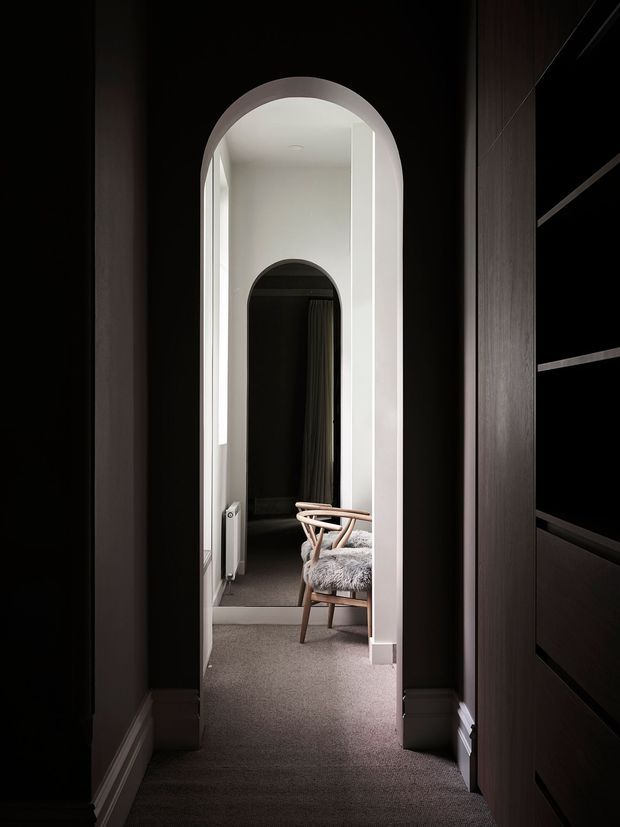 A narrow, moody interior hallway framed by dark built-in joinery and walls, leading through a tall rounded archway into a brighter sitting or dressing area. The corridor has dark carpet, white trim and arch reveals, with a timber-framed chair upholstered in a grey textured fabric visible beyond. A second smaller arched opening and dark curtains create depth at the rear.