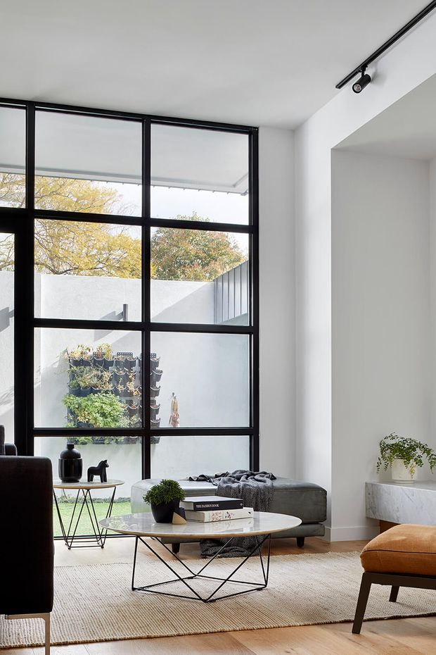 Bright, contemporary living room with a large black-framed steel-look grid window opening to a planted courtyard. The room has white walls, pale timber flooring, a textured neutral rug, a low dark ottoman, sculptural coffee table, black track lighting, and warm cognac leather seating. The overall composition is clean, refined, and light-filled.