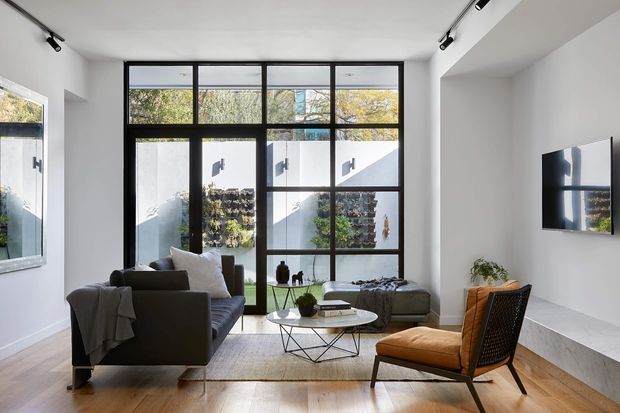 Bright contemporary living room with a dark sofa, sculptural coffee table, upholstered lounge chair, and ottoman arranged on a pale rug over timber flooring. A large black-framed grid window provides abundant natural light and overlooks a planted courtyard, while track lighting and a wall-mounted television complete the clean, gallery-like interior.
