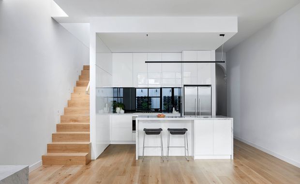 Bright, minimalist open-plan kitchen with a central white island and two bar stools, extensive white cabinetry, integrated stainless-steel appliances, and a black-framed glazed splashback or internal window. Pale timber flooring and an adjacent straight timber staircase create a clean contemporary interior.