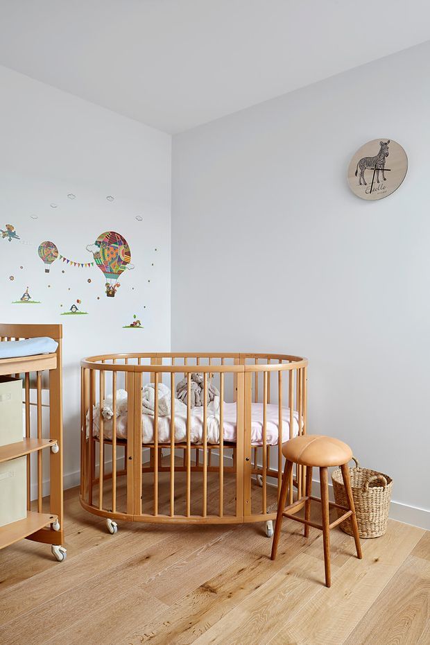 Bright nursery with a round wooden cot as the focal point, a matching timber change table partially visible at left, and a small wooden stool beside a woven basket. The room has pale blue-grey walls, light timber flooring, colourful hot-air-balloon wall decals, and a round animal illustration. Natural light creates a calm, playful atmosphere.