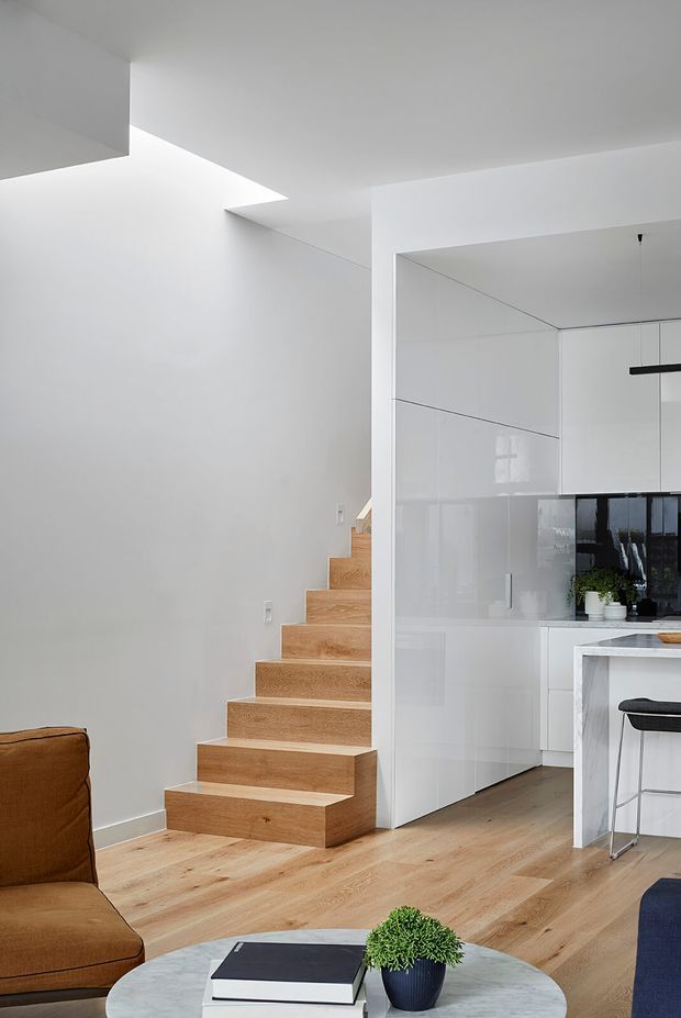 A bright, minimalist open-plan interior showing a light timber staircase beside a sleek white kitchen with integrated cabinetry and a marble-look island. Pale timber flooring continues through the living area, with partial views of a tan sofa and round coffee table. A large skylight or roof opening brings natural light into the stair zone, while a simple black linear pendant hangs over the kitchen island.