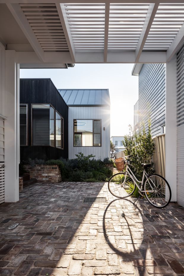 A narrow, sunlit residential courtyard or passage between contemporary buildings. The space has a brick-paved floor, white louvred overhead pergola, horizontal cladding, and a black-and-metal-sided small dwelling or extension at the far end. A bicycle rests against the wall, with planted garden beds and steps near the entrance.
