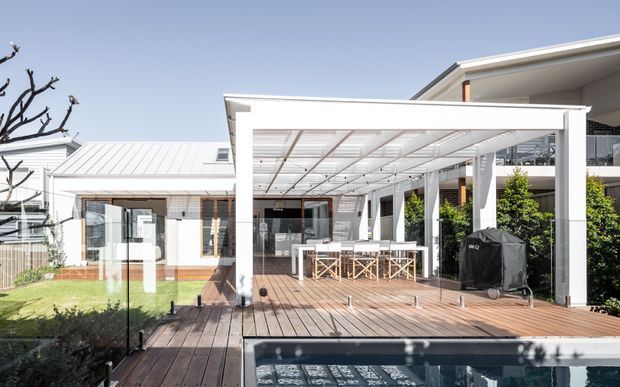 Bright exterior view of a contemporary residential outdoor entertaining area. A timber deck surrounds a swimming pool and leads to a large white-framed pergola with a translucent roof, outdoor dining furniture, barbecue, and glass balustrade. The house has crisp white cladding, metal roofing, extensive glazing, and landscaped garden elements.