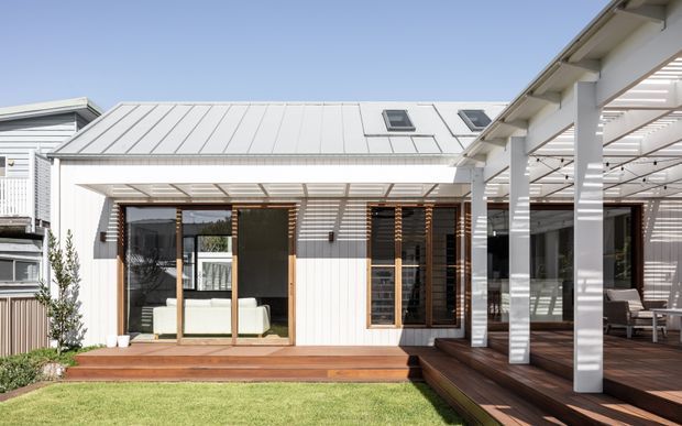 Exterior view of a bright contemporary single-storey home extension with a white standing-seam metal pitched roof, vertical white cladding, extensive glazing, timber screens, and a covered timber deck and pergola. The landscaped lawn and adjoining outdoor entertaining area are visible, with glimpses of the interior through the windows.