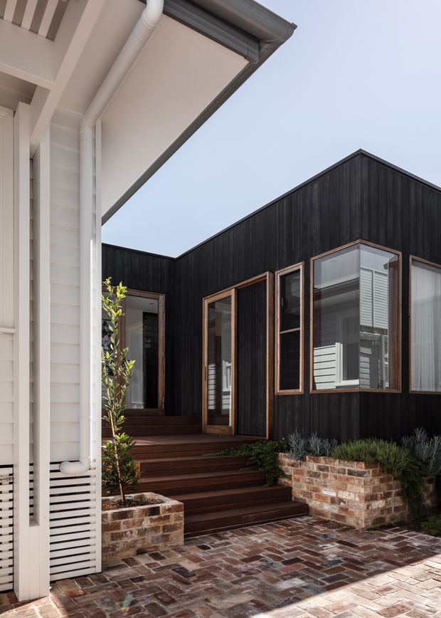 Exterior view of a contemporary residential entry courtyard. A dark, vertically clad gabled extension features timber-framed windows and a glazed door, reached by broad timber steps. The courtyard is paved in reclaimed-looking brick with low brick planter walls and landscaping. A contrasting light weatherboard-clad building with exposed gutters and downpipes frames the left side.