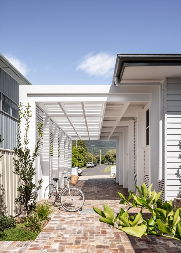 Bright exterior view of a narrow residential passage and entry courtyard framed by white-painted structures. A covered pergola with horizontal slatted roof and side screening leads toward a street and wooded hillside beyond. The path is paved with multicoloured brick or stone pavers, with planted garden beds and a bicycle in the foreground. The adjacent building has light grey weatherboard cladding, white trims, gutters and downpipes, creating a clean contemporary coastal character.
