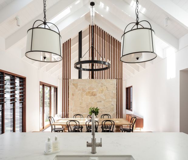 Bright, airy open-plan kitchen and dining area with a large white island and central sink in the foreground. A long timber dining table with black metal chairs sits beyond the island, framed by a tall natural stone feature wall and vertical timber slat screen. The room has a steep white vaulted ceiling with exposed white beams, clerestory-like glazing, dark-framed windows, and three oversized hanging drum and ring pendant lights.