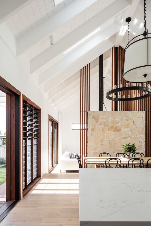 A bright, airy open-plan kitchen and dining space with a dramatic white pitched, timber-lined ceiling and exposed beams. The foreground features a large pale stone kitchen island, while the dining area includes a timber table and black bentwood-style chairs. A rustic natural-stone feature wall, vertical timber screens, dark timber-framed glazing, and large circular black pendant lights add texture and contrast. Sunlight streams through the tall windows and doors onto the light timber floor.